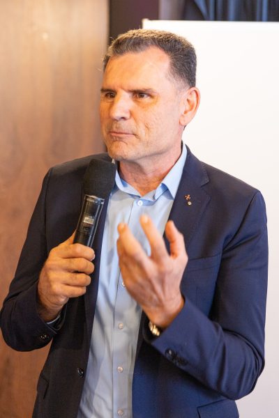 Ein Mann im Anzug hält ein Mikrofon in der Hand und gestikuliert beim Sprechen mit der anderen Hand; er steht in einem Raum in der Nähe einer Weißwandtafel.