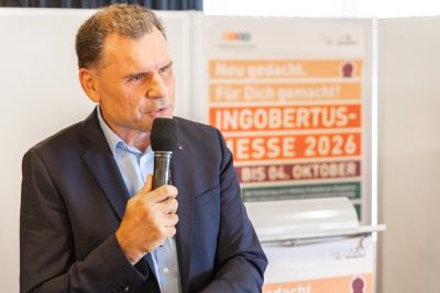 Ein Mann im Anzug spricht in ein Mikrofon. Hinter ihm ist ein buntes Schild mit der Aufschrift INGOBERTUS MESSE 2026 zu sehen, auf dem auch weiterer deutscher Text und Daten zu erkennen sind.