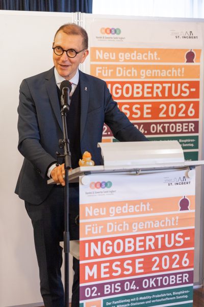 Ein Mann mit Brille und im Anzug spricht an einem Rednerpult, hinter ihm hängen bunte Banner, die für die Ingobertus-Messe 2026 werben, die vom 2. bis 4. Oktober in St. Ingbert stattfindet.
