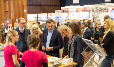 Eine Gruppe von Menschen steht bei einer gut besuchten Indoor-Veranstaltung um einen Stand herum, blättert in Broschüren und unterhält sich; im Hintergrund sind Ausstellungsstücke und andere Besucher zu sehen.