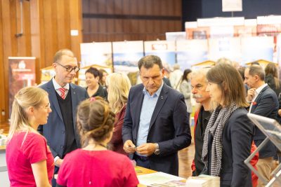 Eine Gruppe von Menschen in Geschäftskleidung unterhält sich mit zwei Frauen in roten Shirts auf einer gut besuchten Veranstaltung oder Konferenz in einem Saal, im Hintergrund sind Infostände und Ausstellungstafeln zu sehen.