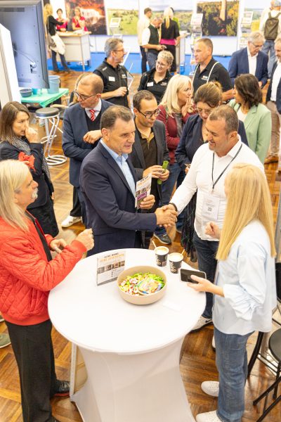 Die Teilnehmer einer Networking-Veranstaltung versammeln sich um einen hohen runden Tisch, geben sich die Hand, unterhalten sich und tauschen Broschüren aus. Auf dem Tisch stehen Kaffeetassen und eine Schale mit Bonbons. Die Atmosphäre ist lebhaft und professionell.