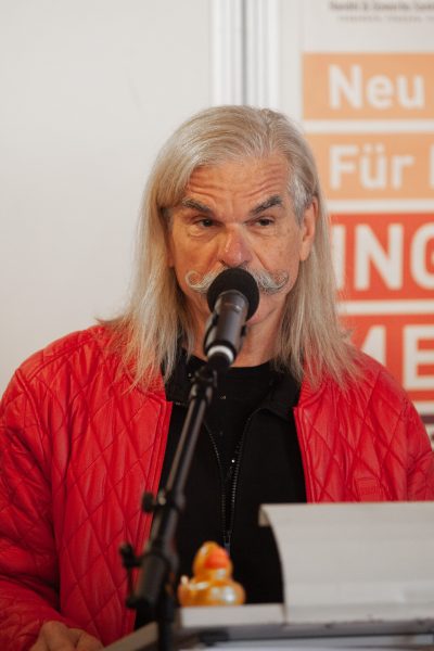 Ein älterer Mann mit langen grauen Haaren und einem gekräuselten Schnurrbart, der eine rote Jacke trägt, spricht in ein Mikrofon. Auf dem Podium vor ihm sitzt eine Gummiente. Im Hintergrund ist ein orange-weißes Schild zu sehen.