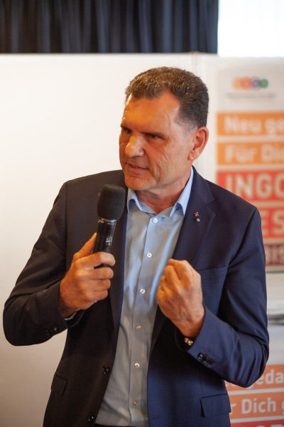 Ein Mann im Anzug spricht in ein Mikrofon und gestikuliert dabei mit der anderen Hand, die zur Faust geballt ist. Er steht in einem Raum vor einem unscharfen Schild mit Schriftzug und scheint sich an ein Publikum zu wenden.
