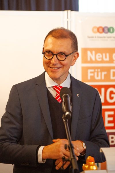 Ein Mann in Anzug und rot gestreifter Krawatte steht lächelnd vor einem Mikrofon. Hinter ihm ist ein unscharfes Schild mit deutschem Text und bunten Logoelementen zu sehen. Die Kulisse wirkt professionell und formell.