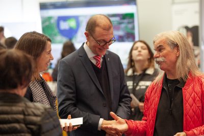 Drei Erwachsene unterhalten sich bei einer Veranstaltung in einem Saal. Ein Mann mit langen grauen Haaren in einer roten Jacke gestikuliert beim Sprechen. Ein weiterer Mann im Anzug und eine Frau, die Unterlagen in der Hand hält, hören zu und lächeln. Im unscharfen Hintergrund sind weitere Personen zu sehen.