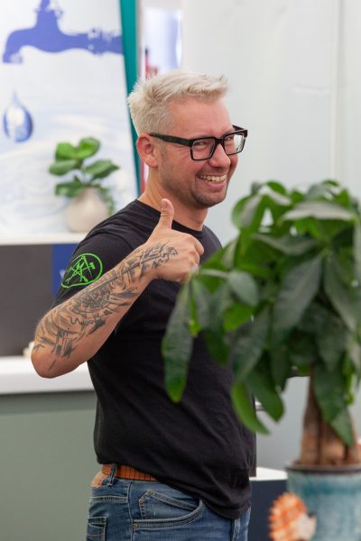 Ein lächelnder Mann mit Brille und Tattoos zeigt den Daumen nach oben in einem Innenraum, im Hintergrund sind Pflanzen und eine blau-weiße Wand zu sehen.
