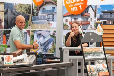 Zwei Personen stehen an einem Stand, an dem Zaunprodukte ausgestellt sind; beide lächeln und zeigen den Daumen nach oben. Im Hintergrund sind Werbeplakate mit Häusern und der Aufschrift Nr. 1 auf Deutsch zu sehen. Broschüren und kleine Fähnchen schmücken den Stand.