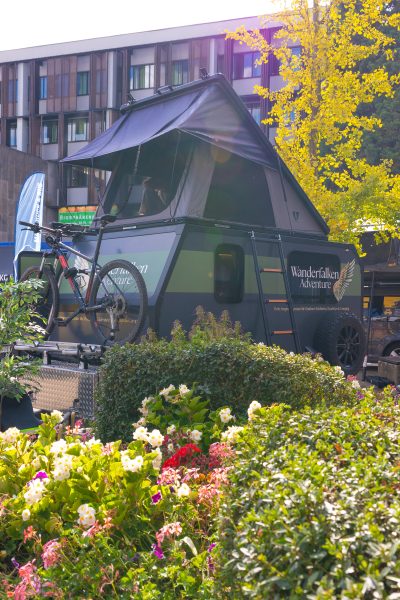 Hinter einem farbenfrohen Blumengarten steht ein Wohnmobil mit einem Dachzelt und einem vorne befestigten Fahrrad – das perfekte Motiv für eine Pressemitteilung über Outdoor-Abenteuer. Im Hintergrund sind ein Gebäude und Bäume zu sehen.