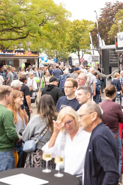Ein gut besuchtes Open-Air-Festival mit Menschen, die sich unterhalten, Imbissständen, Luftballons und Bäumen im Hintergrund. Im Vordergrund stehen zwei Gläser Weißwein auf einem Tisch – eine lebhafte Szene, die sich hervorragend für jede Pressemitteilung eignet.