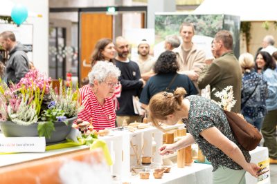 Eine Frau in einem rot gestreiften Hemd steht hinter einem Ausstellungsstand mit Töpferwaren, während eine andere die Ausstellungsstücke begutachtet. Im Hintergrund unterhalten sich die Besucher bei dieser Indoor-Veranstaltung, über die in der aktuellen Pressemitteilung berichtet wird.
