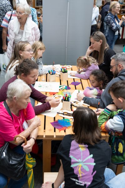 Eine Gruppe von Kindern und Erwachsenen sitzt bei einer gut besuchten Veranstaltung in den Innenräumen um einen Holztisch herum und malt und koloriert mit Filzstiften und Buntstiften, wie in der aktuellen Pressemitteilung zu sehen ist. Auf dem Tisch liegen Malutensilien und bunte Papiere verstreut.