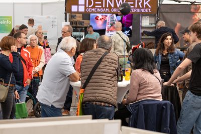 Bei einer gut besuchten Veranstaltung in den Innenräumen steht eine Gruppe von Menschen um einen hohen runden Tisch herum und unterhält sich – Getränke und Taschen in der Hand – unter einem großen DEINWERK-Schild, während die interessierten Teilnehmer eine Pressemitteilung besprechen.