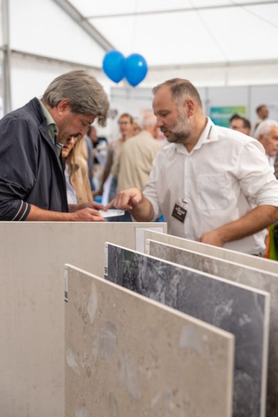 Zwei Männer besprechen bei einer Veranstaltung in einem Saal, umgeben von Menschen und blauen Luftballons, Stein- oder Fliesenmuster, als würden sie sich auf eine Pressemitteilung vorbereiten.