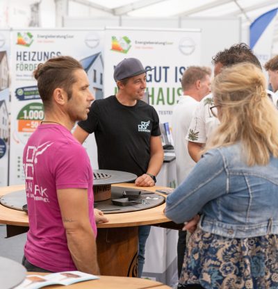 Auf einer Messe stehen Menschen um einen Stand herum und unterhalten sich. Zwischen Ausstellungsplakaten und Broschüren ist eine Pressemitteilung zu sehen, während ein Mann in Schwarz, ein weiterer in Rosa und eine Frau mit Pferdeschwanz anwesend sind.