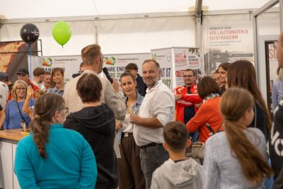 Eine Gruppe von Menschen versammelt sich in einem Raum bei einer Veranstaltung mit Informationsständen und einer lebhaften, geselligen Atmosphäre. Einige lächeln und unterhalten sich, während Luftballons, Plakate und eine Pressemitteilung den lebhaften Hintergrund ergänzen.