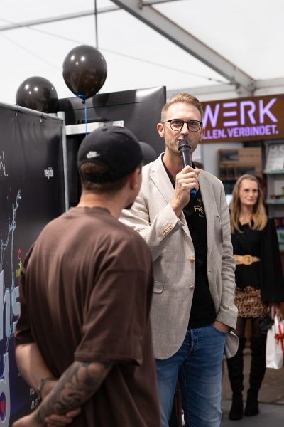 Ein Mann in einem beigen Blazer verliest eine Pressemitteilung in ein Mikrofon, während er einem anderen Mann in einer Mütze und einem braunen Hemd gegenübersteht. Im Hintergrund mischen sich Menschen zwischen Luftballons, wobei Regale und eine Frau, die ein Buch in der Hand hält, zu sehen sind.