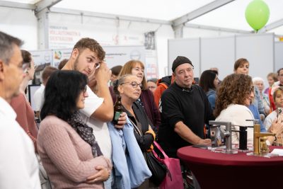 Eine Gruppe von Menschen steht bei einer Veranstaltung in einem Saal dicht beieinander und hört aufmerksam einer Pressemitteilung zu. Einige halten Getränke in der Hand. Ein Mann trägt einen schwarzen Hut, und im Hintergrund schwebt ein grüner Luftballon in der Nähe von Tischen, auf denen verschiedene Gegenstände liegen.