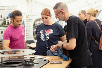 Drei Personen stehen an einem Stand in einer Halle, sehen sich Broschüren und eine Ausstellung an und bereiten möglicherweise eine Pressemitteilung vor. Im Hintergrund sind weitere Personen zu sehen, offenbar auf einer Messe oder Ausstellung.