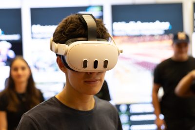 Eine Person, die ein weißes Virtual-Reality-Headset trägt, steht in einem Innenraum, im Hintergrund sind verschwommene Menschen und Bildschirme zu sehen – ganz so, als stamme das Bild aus einer hochmodernen Pressemitteilung.