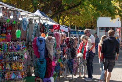 An einem sonnigen Tag stöbern Menschen an einem Marktstand unter Bäumen in Schals und Spielzeug. Eine Pressemitteilung kündigt die Veranstaltung an, während Plüschtiere an einem Gestell hängen und bunte Schals über Ständern drapiert sind.