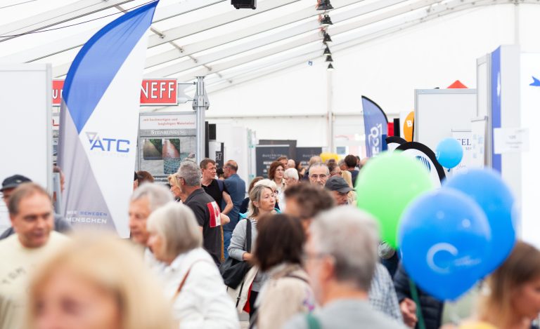 Eine gut besuchte Indoor-Veranstaltung, bei der sich viele Menschen zwischen Ständen und Bannern bewegen; einige halten blaue und grüne Luftballons in der Hand. Die geschäftige Atmosphäre unter dem weißen Zeltdach spiegelt das lebhafte Treiben einer Pressemitteilung wider.