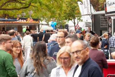 Eine Menschenmenge schlendert an einem bewölkten Tag über einen Markt oder ein Fest im Freien, auf dem es Essensstände, Bäume, Luftballons, Banner und im Hintergrund ein Schild mit der Aufschrift Pressemitteilung zu sehen gibt.