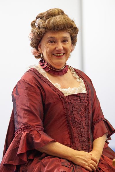 Eine lächelnde Frau in einem aufwendig verzierten, weinroten Kleid mit Spitzenbesatz, Rüschenkragen und einer mit Blumen geschmückten Frisur im Stil der damaligen Zeit, die vor einem schlichten Hintergrund für eine Pressemitteilung posiert.