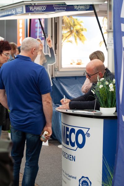 Ein Mann an einem PLAMECO-Stand bespricht eine Pressemitteilung mit einem kahlköpfigen Mitarbeiter, der etwas auf ein Blatt Papier schreibt. In der Nähe stehen mehrere Personen; ein Poster mit einer Strandszene und eine kleine Pflanze schmücken die Theke.