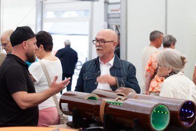 Bei einer Veranstaltung oder Ausstellung in einem Innenraum stehen mehrere Erwachsene um einen Tisch herum und unterhalten sich, während sie zylindrische elektronische Geräte in den Händen halten; die ungezwungene Atmosphäre lässt vermuten, dass gerade eine Pressekonferenz stattfindet. Im Hintergrund sind weitere Personen zu sehen.