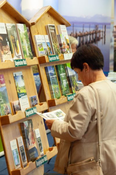 Eine Frau liest eine Broschüre, während sie bei einer Veranstaltung oder Ausstellung in einem Saal vor Holzregalen steht, die mit Reiseprospekten, Reiseführern und Pressemitteilungen gefüllt sind.