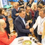Bei einer Networking-Veranstaltung versammelt sich eine Gruppe von Menschen um einen Tisch, auf dem Süßigkeiten, Kaffeetassen, Flyer und eine Pressemitteilung liegen. In der Mitte geben sich zwei Männer die Hand, während andere sich unterhalten und zuschauen.